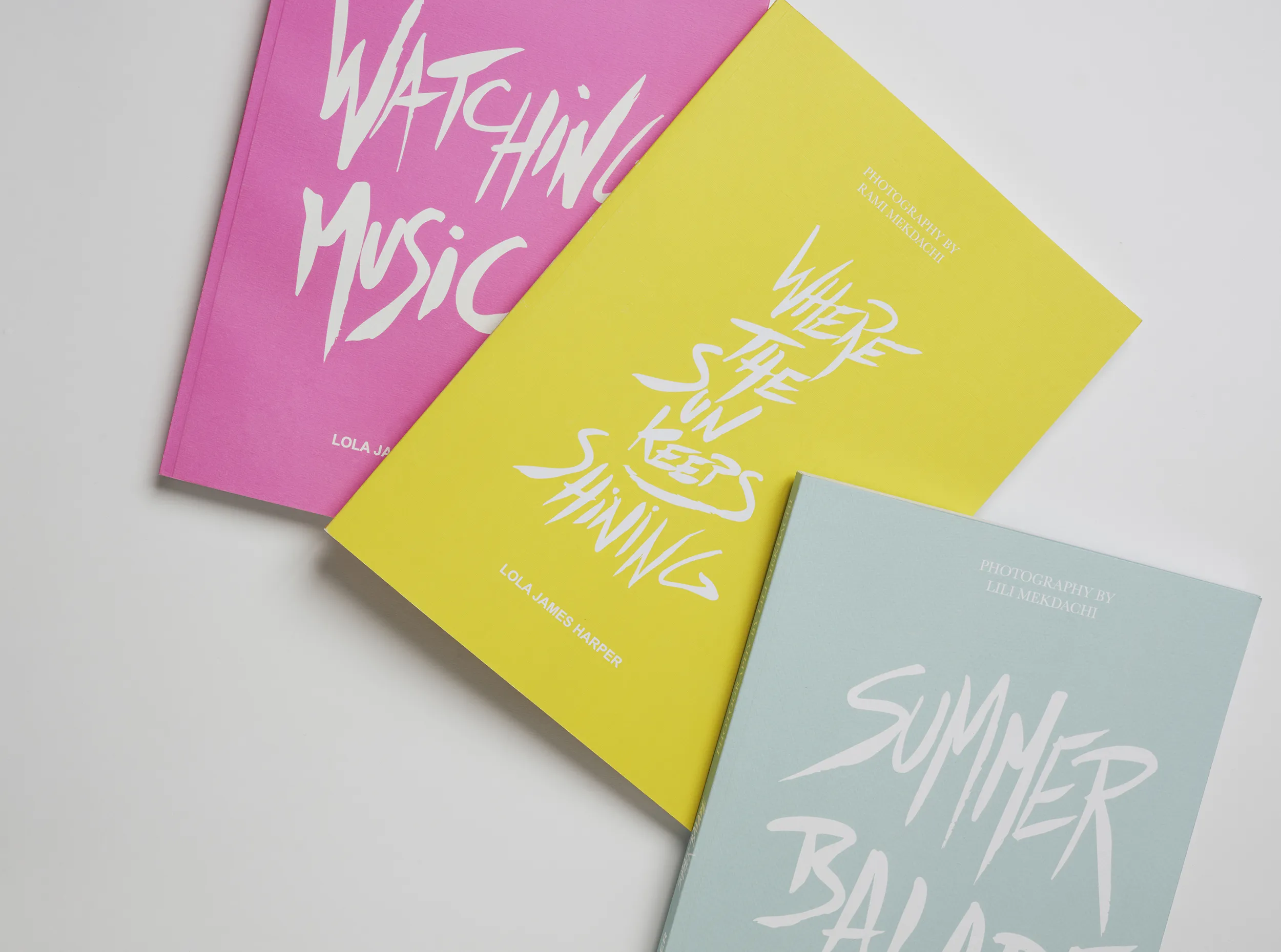 Photo de trois livres posés sur fond blanc : « Watching Music » en rose, « Where the Sun Keeps Shining » en jaune au centre, « Summer Balade » en bleu pâle.